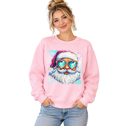 A woman wearing a light pink crewneck sweatshirt with a colorful graphic of a Santa Claus face wearing heart-shaped sunglasses, standing in a decorated holiday kitchen. The brand name 'ORNAMENTALLYYOU' is visible in the bottom right corner.