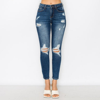 A model is wearing a pair of high-waisted, distressed blue denim jeans with ripped details at the knees and thighs, paired with white heeled sandals against a plain white studio background.