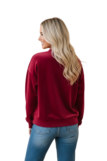 A woman with blonde hair is shown from behind wearing a burgundy crewneck sweatshirt and blue jeans, standing against a solid peach-colored background.