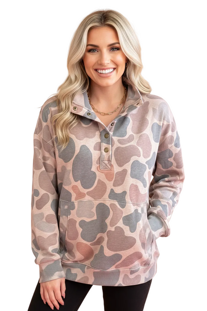 A smiling blonde woman wearing a long-sleeve quarter-placket pullover hoodie with a muted camouflage pattern in dusty pink, taupe, and gray, posed against a solid warm beige background.