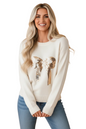 A smiling blonde woman wearing a cream knit sweater with a large sequined bow graphic in metallic gold and rose gold, paired with blue jeans, against a solid peach background.