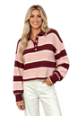 A smiling blonde woman wearing a pink and burgundy striped knit polo sweater with white pants, posed against a solid peach studio background.