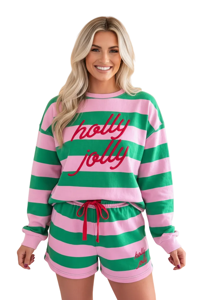 A woman modeling a matching pink and green striped lounge set with red cursive 'holly jolly' text on the sweatshirt and shorts, set against a solid peach background.