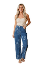 A woman stands against a solid peach studio background wearing blue floral wide-leg jeans, a cream lace camisole, and metallic sandals.