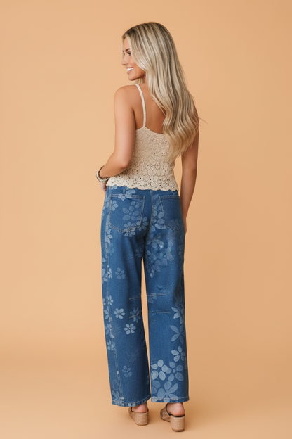 A woman is shown from the back wearing wide-leg blue jeans with a light blue floral pattern, paired with a beige crochet camisole and beige heels, set against a solid peach studio background.