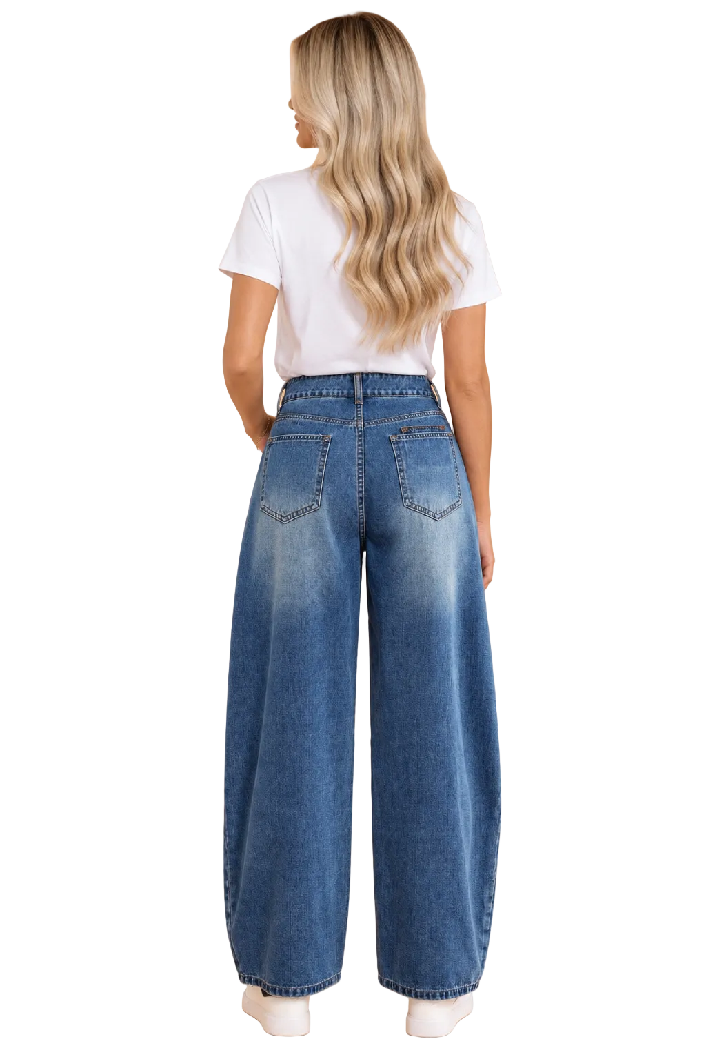A woman viewed from behind wearing high-waisted, wide-leg blue denim jeans, a white t-shirt, and white sneakers against a solid peach studio background.
