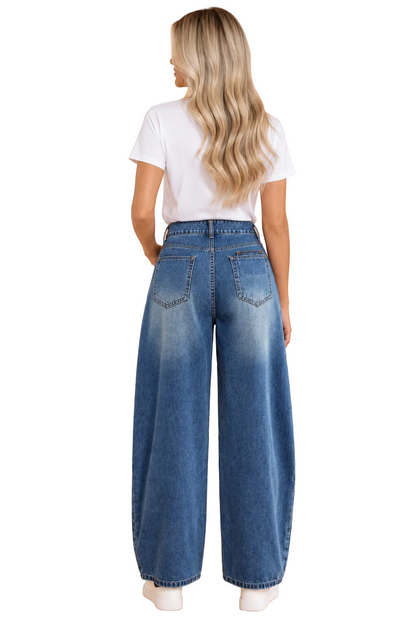 A woman viewed from behind wearing high-waisted, wide-leg blue denim jeans, a white t-shirt, and white sneakers against a solid peach studio background.