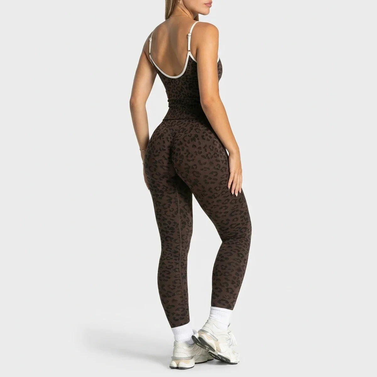 A woman modeling a brown leopard-print athletic jumpsuit from a rear three-quarter angle, wearing white sneakers against a light gray studio background.