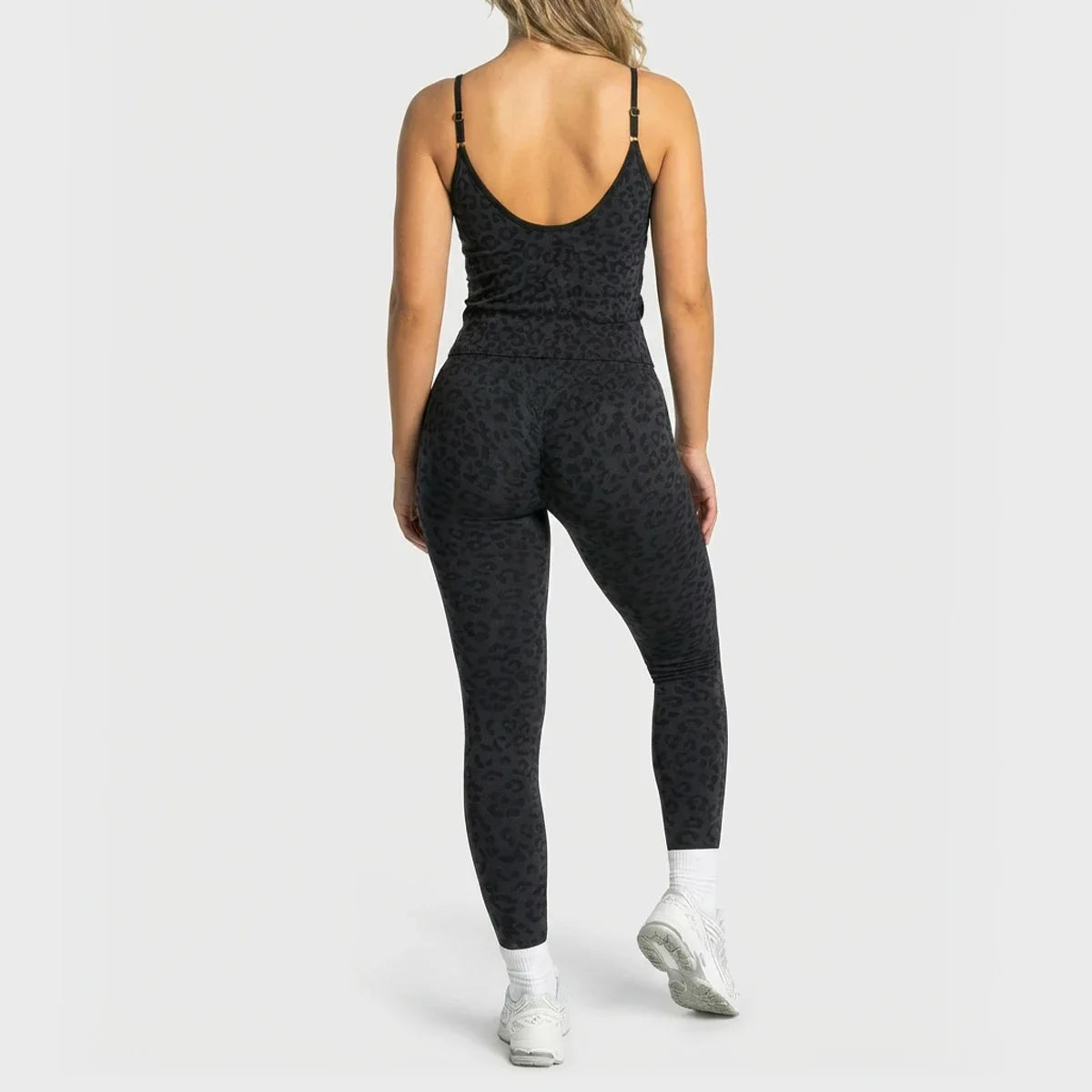 A woman shown from the back wearing a black leopard-print activewear set, consisting of a spaghetti-strap tank top and matching high-waisted leggings, paired with white socks and white sneakers, against a light gray studio background.