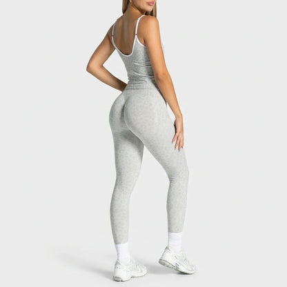 A woman modeling a light gray heathered athletic jumpsuit from a rear three-quarter angle, wearing white socks and white sneakers against a light gray studio background.
