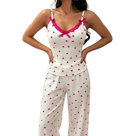 A woman modeling a white ribbed two-piece pajama set with a red heart print, featuring a camisole top with pink lace trim and matching pants, set against a bedroom background.