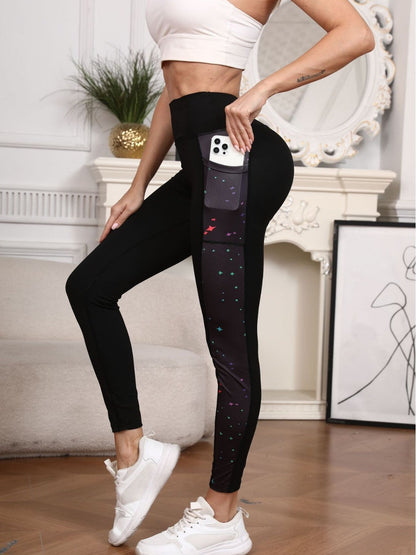 Black high-waisted leggings with a side pocket containing a phone, featuring a colorful galaxy print on the lower leg, modeled in a bright indoor setting.