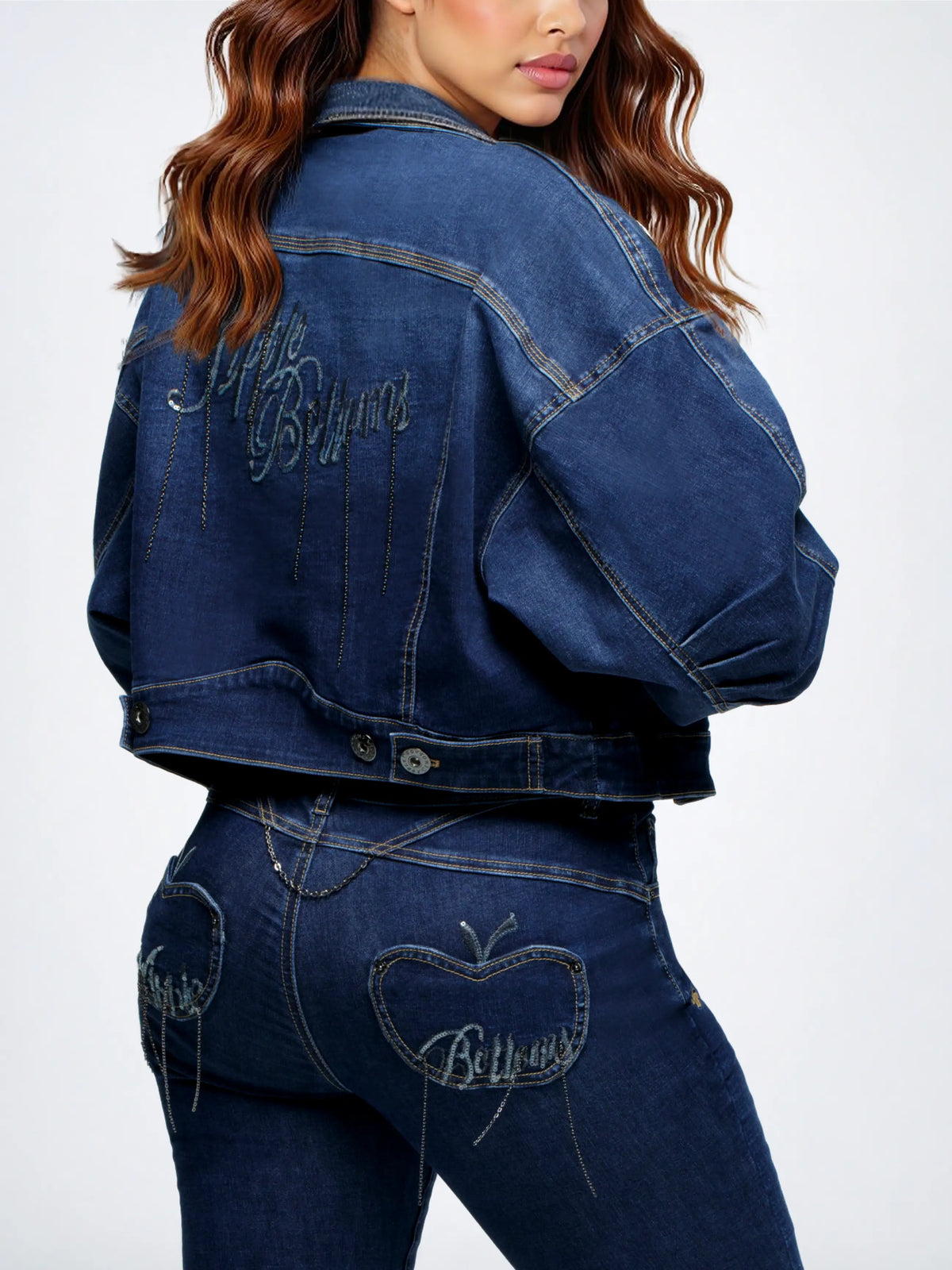 A model shown from the back wearing a dark blue cropped denim jacket and matching denim jeans, both featuring 'Billionaire' embroidered in silver on the back pocket.