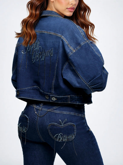 A model shown from the back wearing a dark blue cropped denim jacket and matching denim jeans, both featuring 'Billionaire' embroidered in silver on the back pocket.