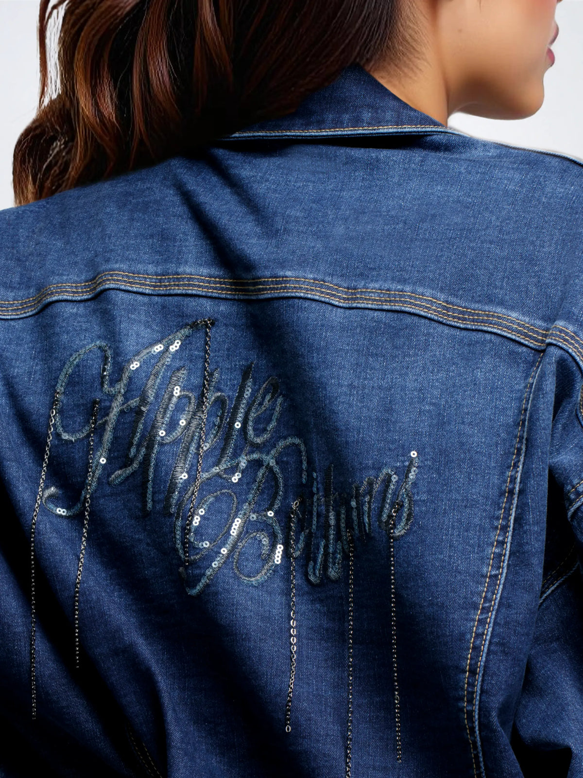 Close-up back view of a person wearing a blue denim jacket with large sequined script reading 'Nipple Bottoms' and dangling chain embellishments.