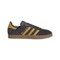 A black suede Adidas Gazelle sneaker with gold stripes, a gold heel tab, and gold 'MADE IN GERMANY' text, paired with a brown gum sole, shown in a side profile on a white background.