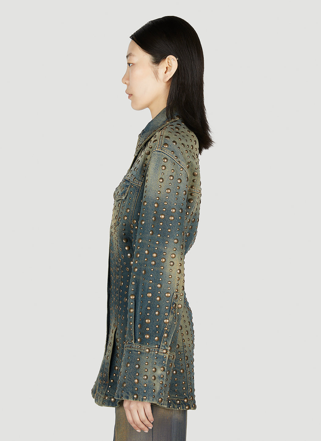 A model shown in profile wearing a dark denim jacket covered in gold-tone studs, set against a solid light gray background.
