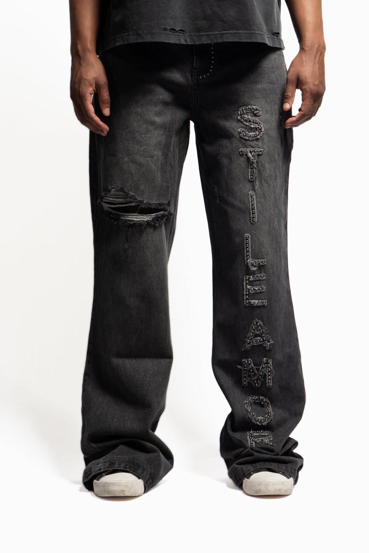 A person wearing very baggy black washed denim jeans with distressed rips on the knees and large vertical embroidered text down one leg, shown against a plain light background.
