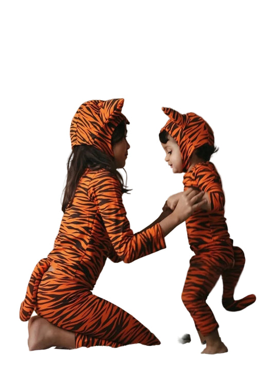 A woman and a young child wearing matching orange and black tiger-print hooded pajamas pose together on a bed with white bedding, a small teddy bear, and soft neutral decor in the background.
