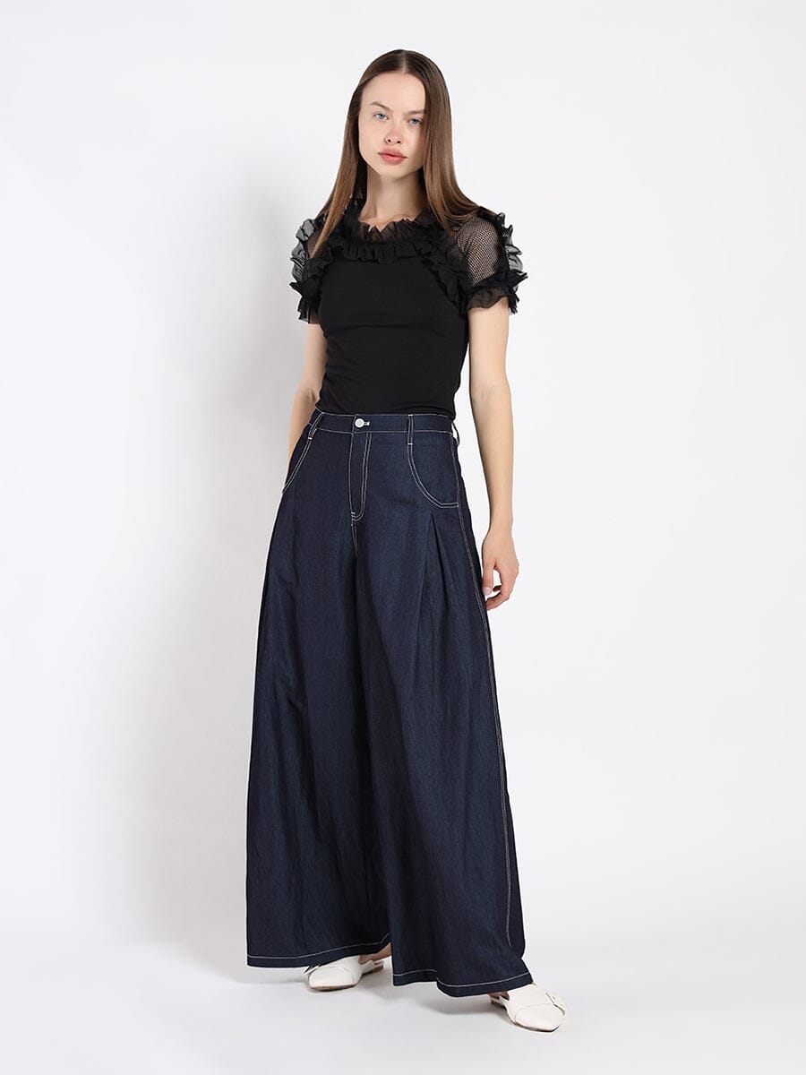 Woman wearing a black top with sheer sleeves and dark blue wide-leg pants on a white background