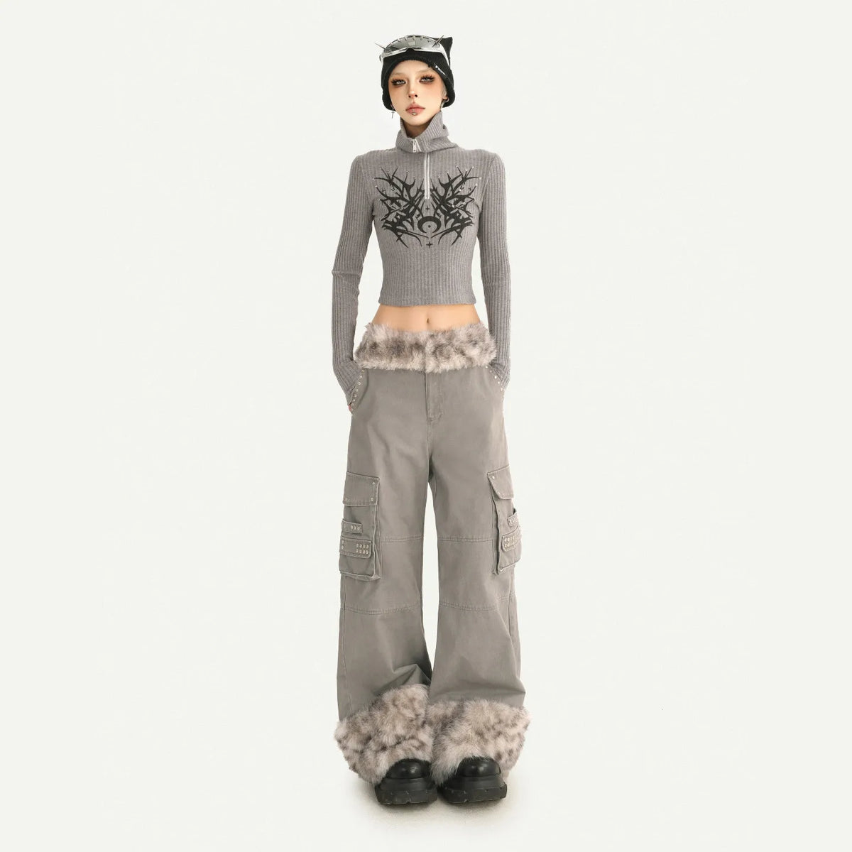 A model stands against a light gray studio background wearing a gray ribbed mock-neck crop top with a black graphic print, matching gray wide-leg cargo pants with faux-fur trim at the hem, a black beanie with cat ears, and black shoes.