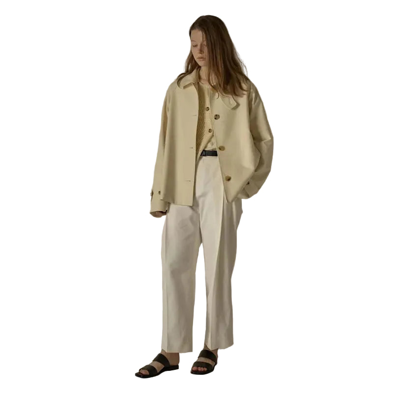 A model stands in a studio wearing a cream-colored double-breasted jacket over a matching top, paired with white wide-leg trousers and black slide sandals.